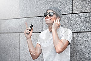 Cheerful guy dressed in gray t-shirt, sunglasses and hat at the street, listening to music with earphones, holding mobile phone