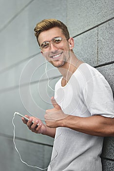 Cheerful guy dressed in white t-shirt at the street, listening to music with earphones, holding mobile phone.