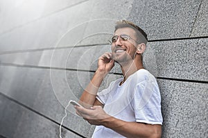 Cheerful guy dressed in white t-shirt at the street, listening to music with earphones, holding mobile phone.