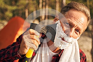 Cheerful guy shaving beard with axe in nature