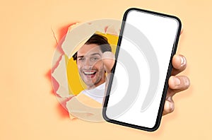 Cheerful guy showing white empty smartphone screen through torn paper