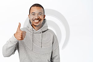 Cheerful guy shows thumbs up, likes and approves smth, recommends company or website, stands over white background