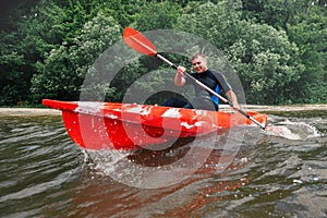 Cheerful guy in wetsuit sits in a red kayak and row with a pad