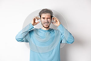 Cheerful guy wink at camera and smiling, listening music in wireless headphones, white background