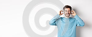 Cheerful guy wink at camera and smiling, listening music in wireless headphones, white background