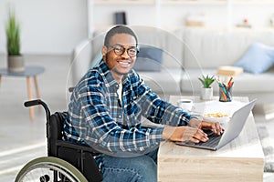 Cheerful handicapped black man in wheelchair using laptop for online job from home, having business meeting on web