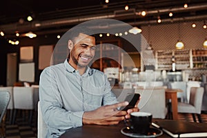 Cheerful handsome african young man relaxing in modern cafe using mobile phone while drinking coffee