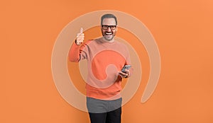 Cheerful handsome entrepreneur holding mobile phone and gesturing thumbs up on orange background