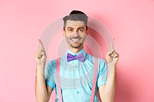Cheerful handsome guy smiling at camera, pointing fingers up, showing advertisement on top of pink background