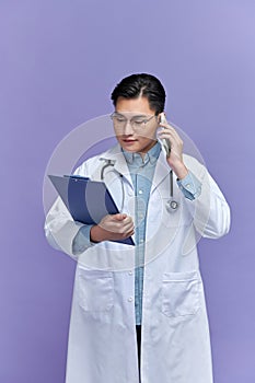 Cheerful handsome male doctor holding clipboard and talking on cell phone on purple background