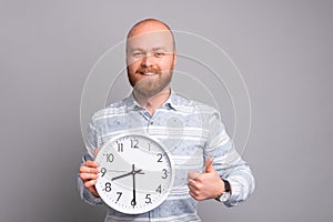 Cheerful handsome man with beard holding white clock and showing thumb up