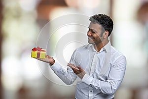 Cheerful handsome man presenting gift box on blurred background.