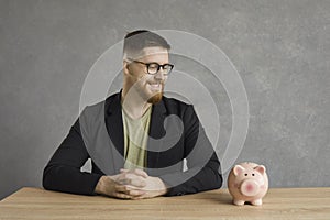 Cheerful handsome man sitting at the table and looking at the piggy bank with a smile on his face.