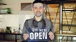 Cheerful handsome man small coffee-house owner is holding `we are open` sign while standing inside coffee shop. Opening