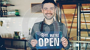 Cheerful handsome man small coffee-house owner is holding We Are Open sign while standing inside coffee shop. Opening