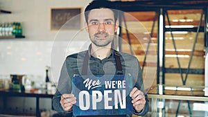 Cheerful handsome man small coffee-house owner is holding We Are Open sign while standing inside coffee shop. Opening