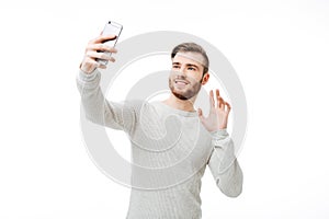 Cheerful handsome man taking selfie and waving hello to the front camera over white background