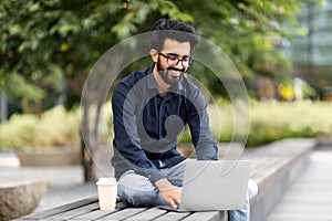 Cheerful millennial indian guy freelancer working on street, using laptop