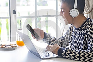 Cheerful handsome teenager using laptop at home in front of the window wearing headphones and holding smart phone. Millenial