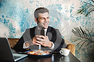 Cheerful handsome young businessman sitting in cafe using laptop computer have a breakfast or dinner eat using mobile phone