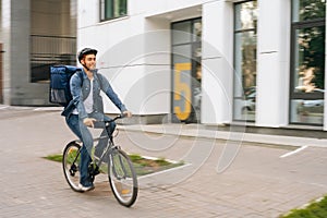 Cheerful handsome young delivery man with thermo backpack riding bicycle in city street on blurred background of office