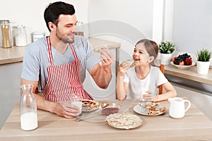 Cheerful handsome young male and little child eat pancakes together, drinks fresh milk, enjoy breakfast at kitchen