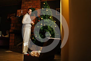 Cheerful handsome young man in cozy sweater decorating Christmas tree with colorful shiny balls near fireplace in dark