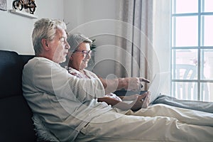 Cheerful happy beautiful caucasian old aged senior couple sit down and relax on the soa at home looking and using a personal