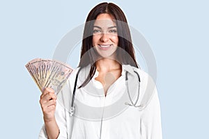 Cheerful happy brunette female doctor in white robe, holds cash, earns salary by curing patients, stands against blue background.