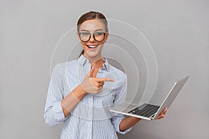 Cheerful happy business young woman posing isolated over grey wall background using laptop computer pointing.