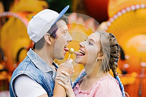 A cheerful happy couple of a guy and a girl eat candy on a stick, smile and stick out their tongues to a Lollipop