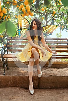 Cheerful happy fairy girl sits on bench in fabulous lemon garden. Fantasy woman in yellow dress pixie costume, fake