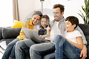 Cheerful happy family of four using laptop sitting on the sofa in cozy apartment