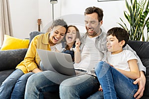 Cheerful happy family of four using laptop sitting on the sofa in cozy apartment, parents and two kids watching movie on