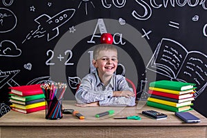 Cheerful and happy hellion schoolboy sitting at the desk with books, school supplies, with a red apple on the top of his