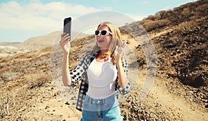 Cheerful happy laughing young woman taking selfie with mobile phone or talking on video call waving hand greeting on a hiking