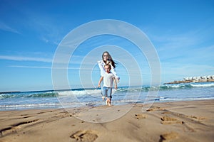 Cheerful happy mom catches her daughter while running barefoot, playing together on beach, leaving footsteps on wet sand