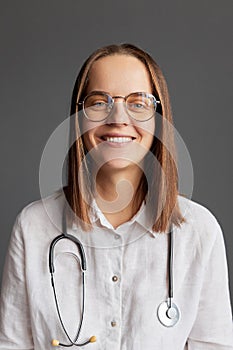 Cheerful happy professional woman doctor in glasses and white medical lab coat looking at camera sitting at workplace in clinic