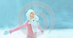 Cheerful happy smiling young woman on winter day wearing hat, sweater and scarf on snowy forest background with snowflakes