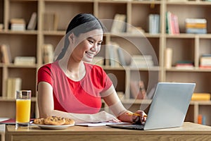 Cheerful happy young asian lady student watch online lesson on laptop, chatting and have breakfast