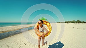 Cheerful happy young attractive woman runs on empty sunny sandy beach holding yellow inflatable pineapple floating ring