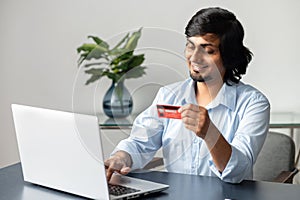 Cheerful and happy young indian man with credit card in hands making purchases online