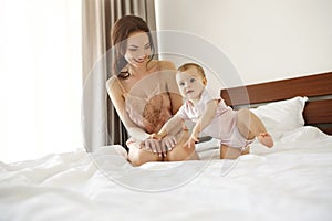 Cheerful happy young mom in sleepwear and her baby daughter smiling playing sitting on bed in morning.