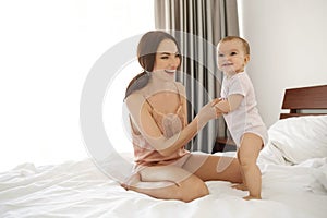 Cheerful happy young mom in sleepwear and her baby daughter smiling playing sitting on bed in morning.