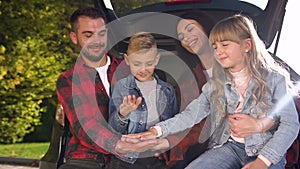 Cheerful happy young parents with their teen children sitting in the trunk and putting hands on one another before car