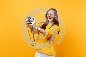 Cheerful happy young woman in heart eyeglasses doing taking selfie shot on retro vintage photo camera isolated on bright
