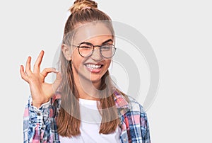 Cheerful happy young woman smiling broadly, dressed in white t-shirt, plaid shirt and round transparent eyewear, shows okay