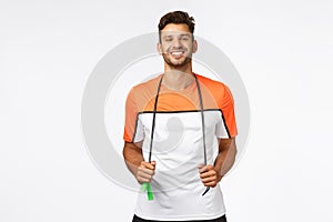 Cheerful, healthy handsome man in sportswear holding jumping rope around neck, smiling pleased and enthusiastic after