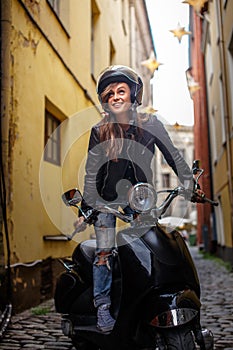 Cheerful hipster girl wearing a leather jacket and ripped jeans standing in a black classic scooter on the old narrow