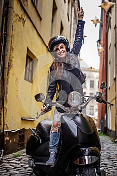 Cheerful hipster girl wearing a leather jacket and ripped jeans standing in a black classic scooter on the old narrow